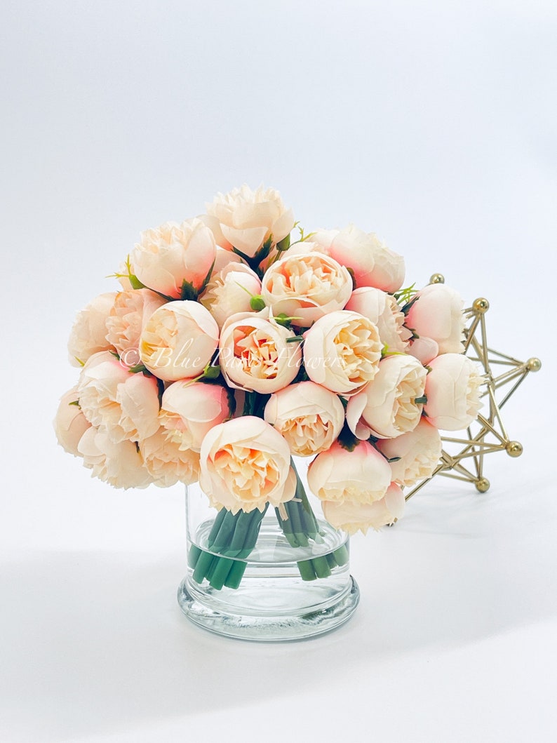 May include: A bouquet of blush pink artificial peonies arranged in a clear glass vase. The vase has a thick base and is sitting on a white surface.