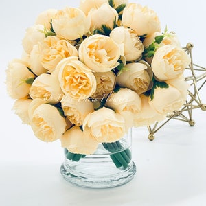 May include: A bouquet of cream-colored artificial peonies in a clear glass vase. The flowers are arranged in a circular shape and have a realistic appearance.