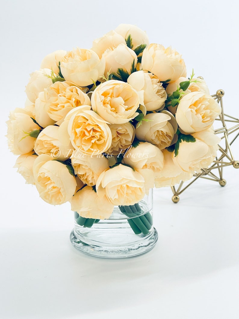 May include: A bouquet of cream-colored artificial peonies in a clear glass vase. The flowers are arranged in a circular shape and have a realistic appearance.