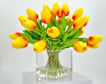 May include: A vibrant display of yellow and red tulips with green foliage, presented in a clear, square glass vase. The vase sits on a white surface. The text "Blue Paris Flowers" is visible. The arrangement is a bright, colourful floral display.