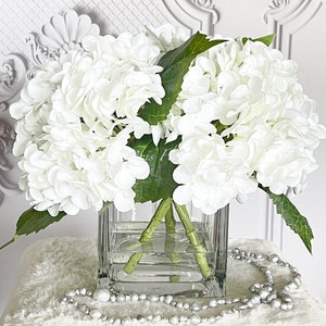 May include: A square glass vase filled with white hydrangea flowers and green leaves. The vase sits on a white fluffy surface, with a strand of silver beads draped in front. The background is a white wall with ornate molding.