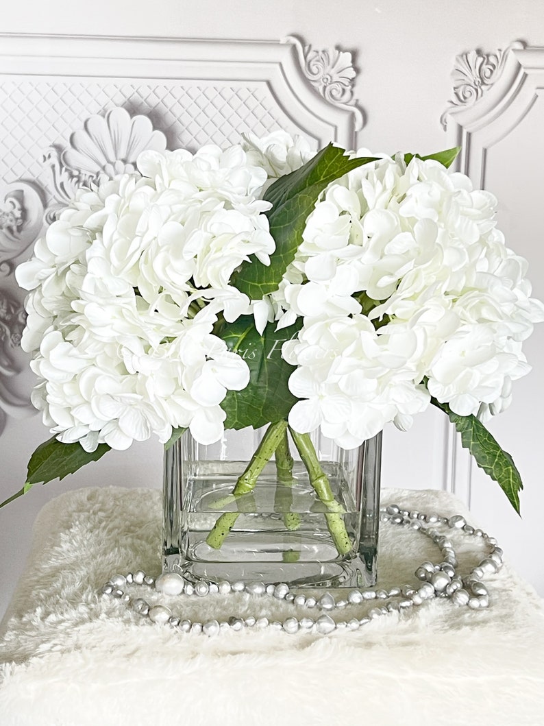 May include: A square glass vase filled with white hydrangea flowers and green leaves. The vase sits on a white fluffy surface, with a strand of silver beads draped in front. The background is a white wall with ornate molding.