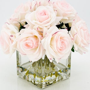 May include: A bouquet of pale pink roses arranged in a clear square glass vase. The roses have a white base and pink edges.