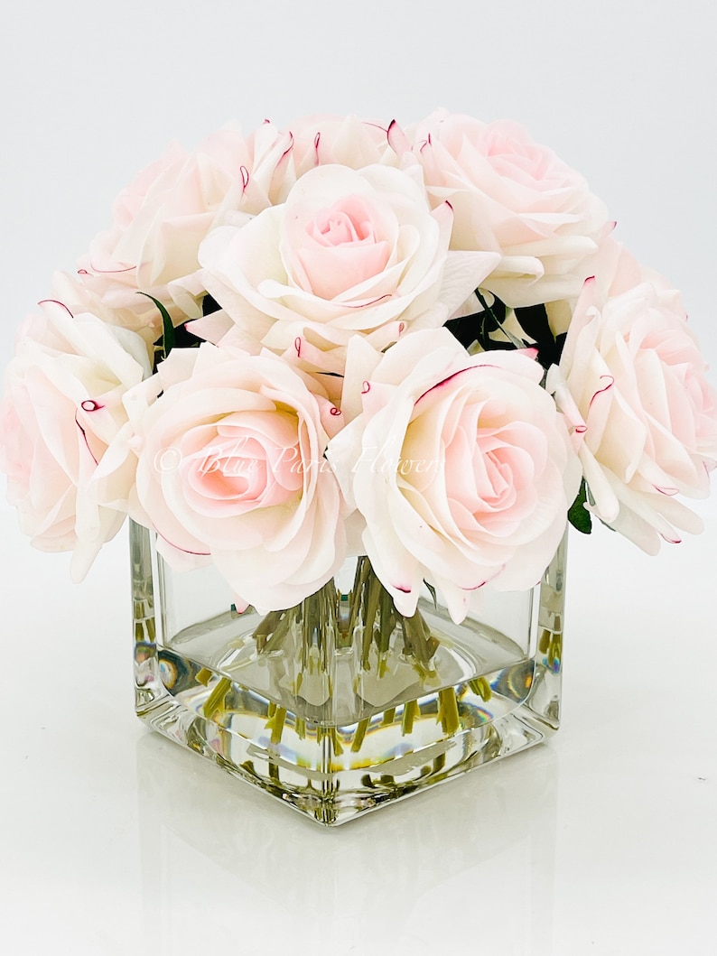 May include: A bouquet of pale pink roses arranged in a clear square glass vase. The roses have a white base and pink edges.