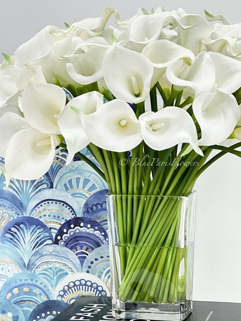 May include: A bouquet of white calla lilies arranged in a clear glass vase. The vase is filled with water and the stems of the flowers are visible. The flowers are in a room with a blue and gold patterned wallpaper.