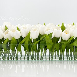 May include: A rectangular glass vase filled with white tulips with green leaves. The vase is sitting on a white surface.