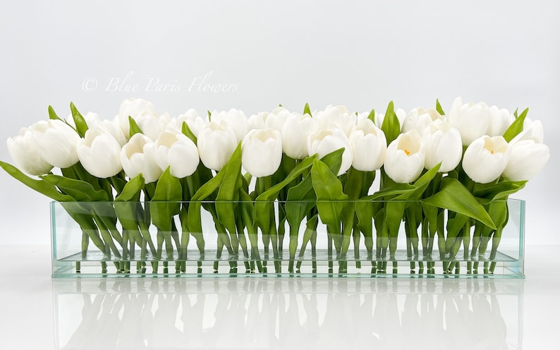 May include: A rectangular glass vase filled with white tulips with green leaves. The vase is sitting on a white surface.