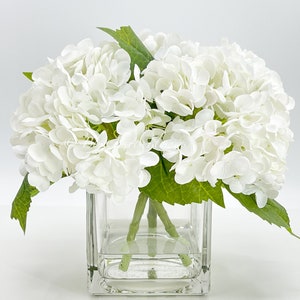 May include: A bouquet of white hydrangea flowers with green leaves in a clear, square glass vase. The flowers are clustered together, creating a full, rounded shape. The vase is filled with water, and the arrangement is set against a white background.