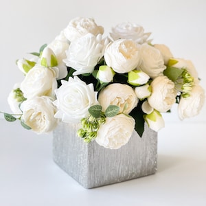 May include: A bouquet of white peonies and roses arranged in a silver square vase.