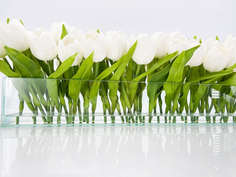 May include: A glass vase filled with white tulips with green leaves. The vase is clear and rectangular.