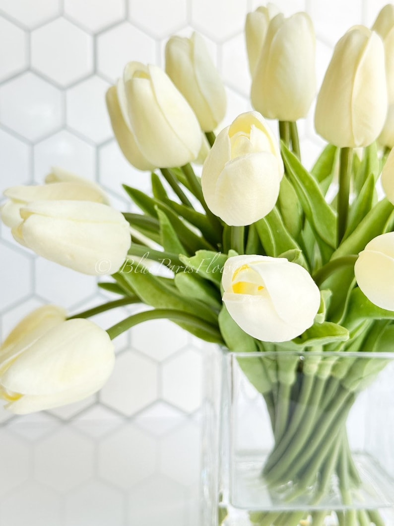 May include: A bouquet of white tulips arranged in a clear glass vase. The tulips are in a simple, modern style and would make a beautiful centerpiece for any home.