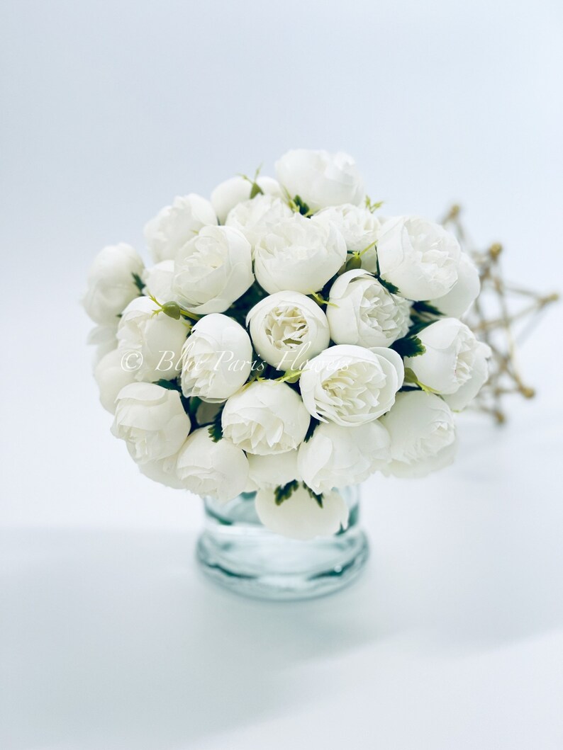 Pu&ograve; includere: Un bouquet di peonie artificiali bianche in un vaso di vetro trasparente. I fiori sono disposti a grappolo e hanno un aspetto realistico.