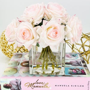 May include: A bouquet of pale pink silk roses arranged in a clear glass cube vase. The vase is sitting on top of two books, one with the title "Love,  Manuela" and the other with the title "Herm&eacute;s Macaron".