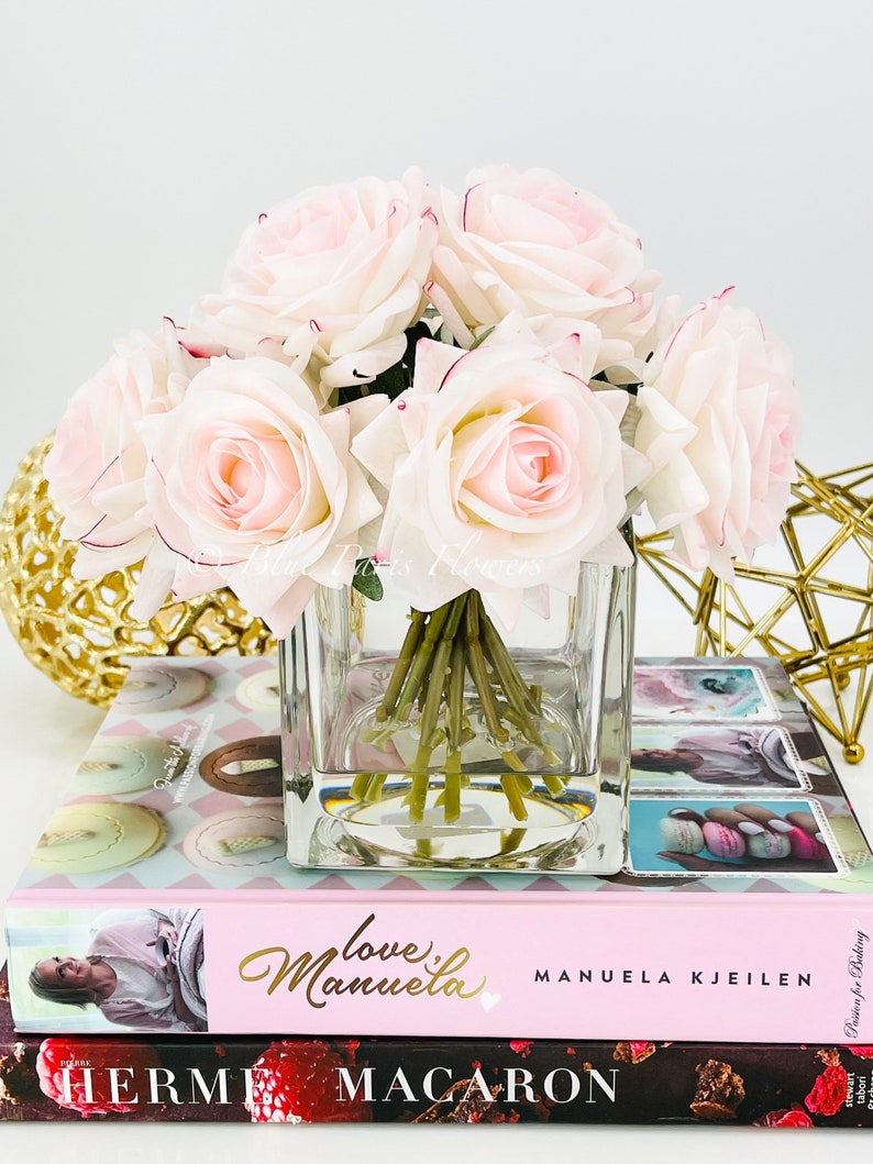 May include: A bouquet of pale pink silk roses arranged in a clear glass cube vase. The vase is sitting on top of two books, one with the title "Love,  Manuela" and the other with the title "Herm&eacute;s Macaron".
