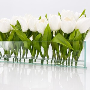 May include: A glass vase filled with white tulips and green leaves. The vase is clear and rectangular.