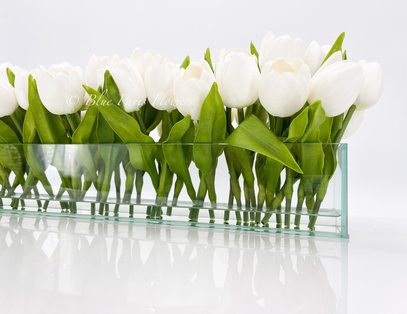 May include: A glass vase filled with white tulips and green leaves. The vase is clear and rectangular.