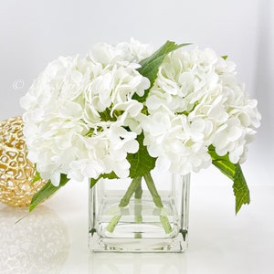 May include: A bouquet of white hydrangea flowers with green leaves in a clear, square glass vase. A gold decorative sphere with a textured surface sits to the left of the vase. The arrangement is on a white surface.