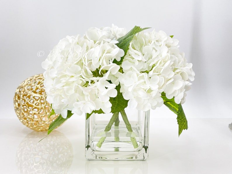 May include: A bouquet of white hydrangea flowers with green leaves in a clear, square glass vase. A gold decorative sphere with a textured surface sits to the left of the vase. The arrangement is on a white surface.