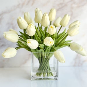 Op de afbeelding: Een boeket crèmekleurige kunstmatige tulpen gerangschikt in een transparante glazen kubusvaas. De tulpen zijn in een dichte groep met groene stelen en bladeren.