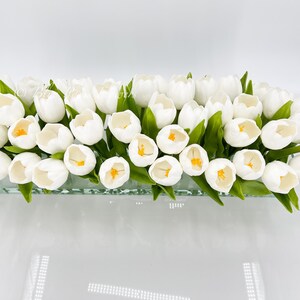 May include: A glass vase filled with a bouquet of white tulips with yellow centers. The vase is clear and has a textured surface.