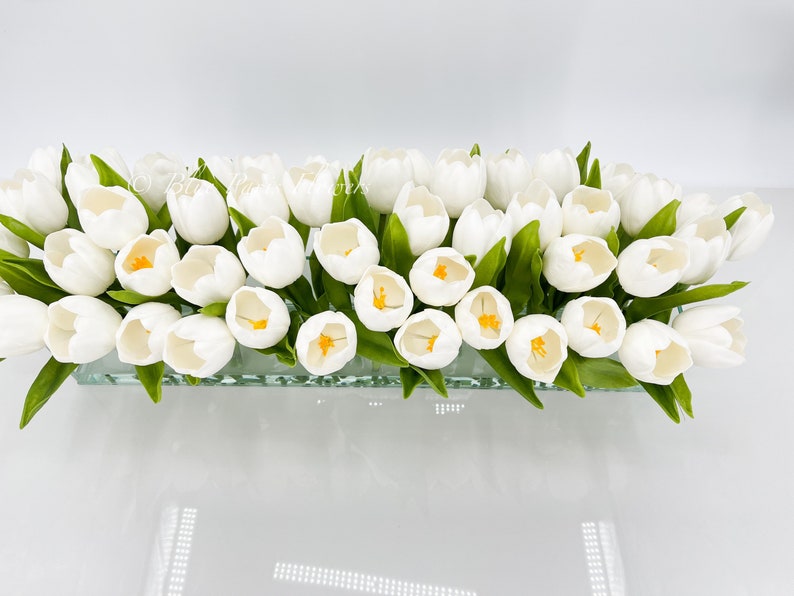 May include: A glass vase filled with a bouquet of white tulips with yellow centers. The vase is clear and has a textured surface.