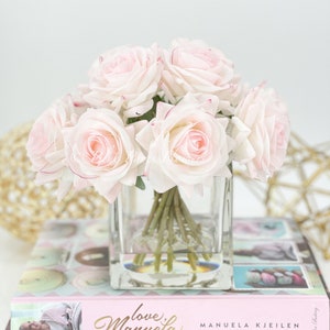 May include: A bouquet of pale pink roses in a clear glass vase. The vase is sitting on top of a pink and white book with the title "Love,  Manuela" and the author "Manuela Kjeilen".
