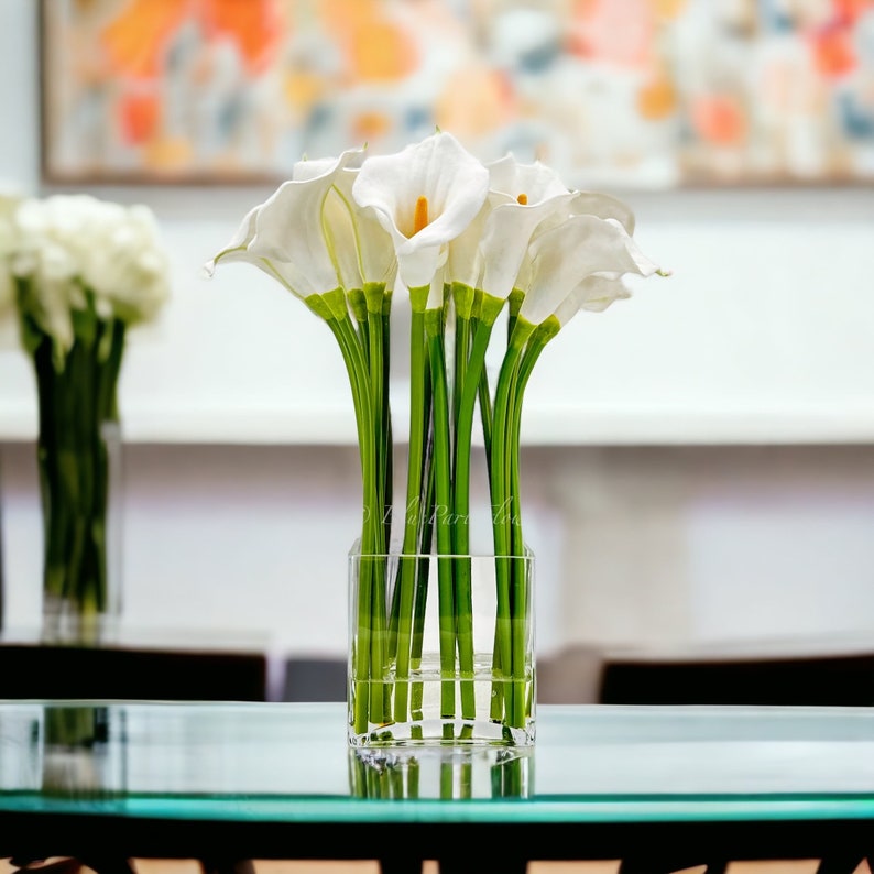 May include: A bouquet of white calla lilies arranged in a clear glass vase. The vase is sitting on a glass table.