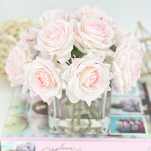 May include: A bouquet of pale pink roses with a hint of red at the edges of the petals, arranged in a clear glass vase. The roses are sitting on a stack of books with a pink cover.