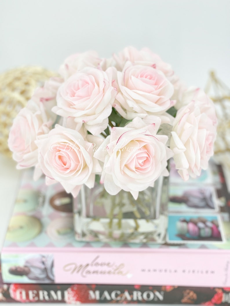May include: A bouquet of pale pink roses with a hint of red at the edges of the petals, arranged in a clear glass vase. The roses are sitting on a stack of books with a pink cover.