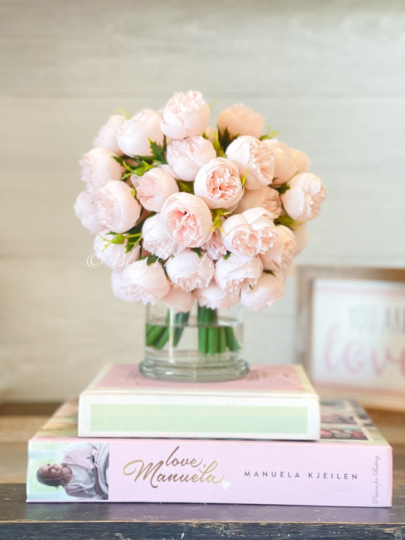 May include: A bouquet of pale pink peonies in a clear glass vase, sitting on top of two books. The top book is white with a green band and the bottom book is pink with the title "love manuela" and the author "MANUELA KJEILEN".