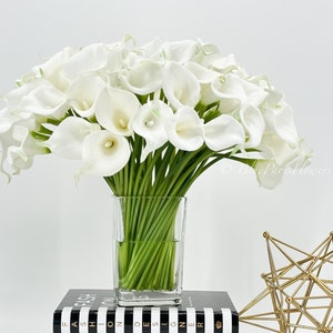May include: A bouquet of white calla lilies arranged in a clear glass vase. The vase is sitting on a black and white striped book.