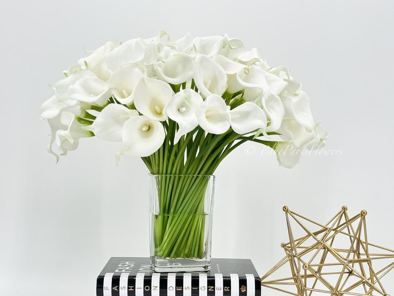 May include: A bouquet of white calla lilies arranged in a clear glass vase. The vase is sitting on a black and white striped book.