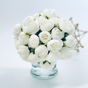 May include: A bouquet of white artificial peonies in a clear glass vase. The flowers are arranged in a cluster and have a realistic appearance.