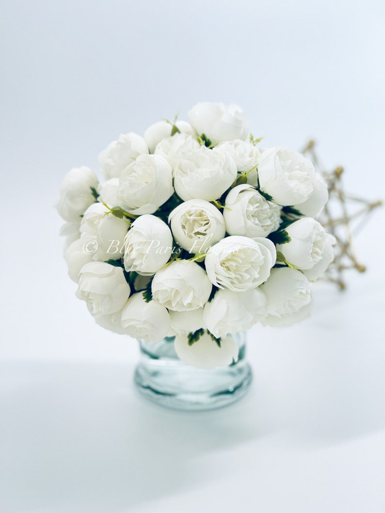 May include: A bouquet of white artificial peonies in a clear glass vase. The flowers are arranged in a cluster and have a realistic appearance.