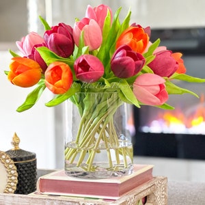 May include: A bouquet of pink, orange and red tulips in a clear glass vase. The vase is sitting on a stack of books on a wooden table.