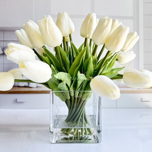 May include: A bouquet of white artificial tulips arranged in a clear glass square vase. The vase is sitting on a white surface.