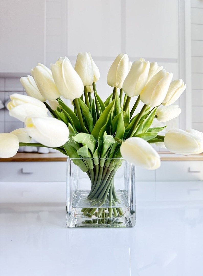 May include: A bouquet of white artificial tulips arranged in a clear glass square vase. The vase is sitting on a white surface.