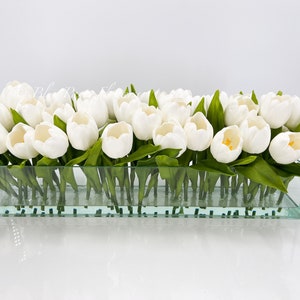 May include: A glass vase filled with white artificial tulips. The vase is clear and rectangular.