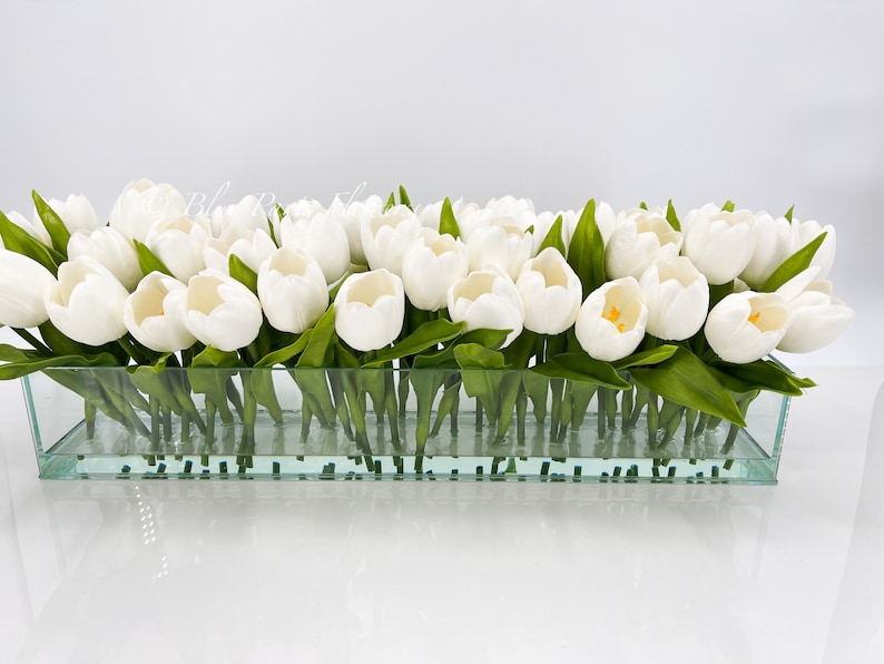 May include: A glass vase filled with white artificial tulips. The vase is clear and rectangular.