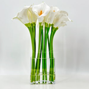 May include: A bouquet of white calla lilies arranged in a clear glass cube vase. The lilies have green stems and are arranged in a symmetrical pattern.