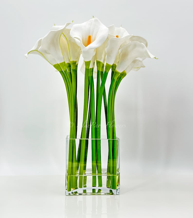 May include: A bouquet of white calla lilies arranged in a clear glass cube vase. The lilies have green stems and are arranged in a symmetrical pattern.