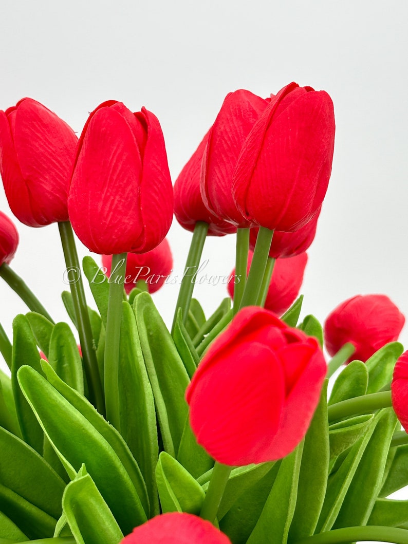 May include: A bouquet of red artificial tulips with green stems and leaves. The tulips are arranged in a cluster, with the blooms facing outward.