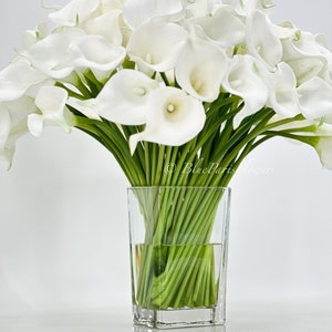 May include: A bouquet of white calla lilies arranged in a clear glass vase. The lilies are in full bloom and have long green stems.