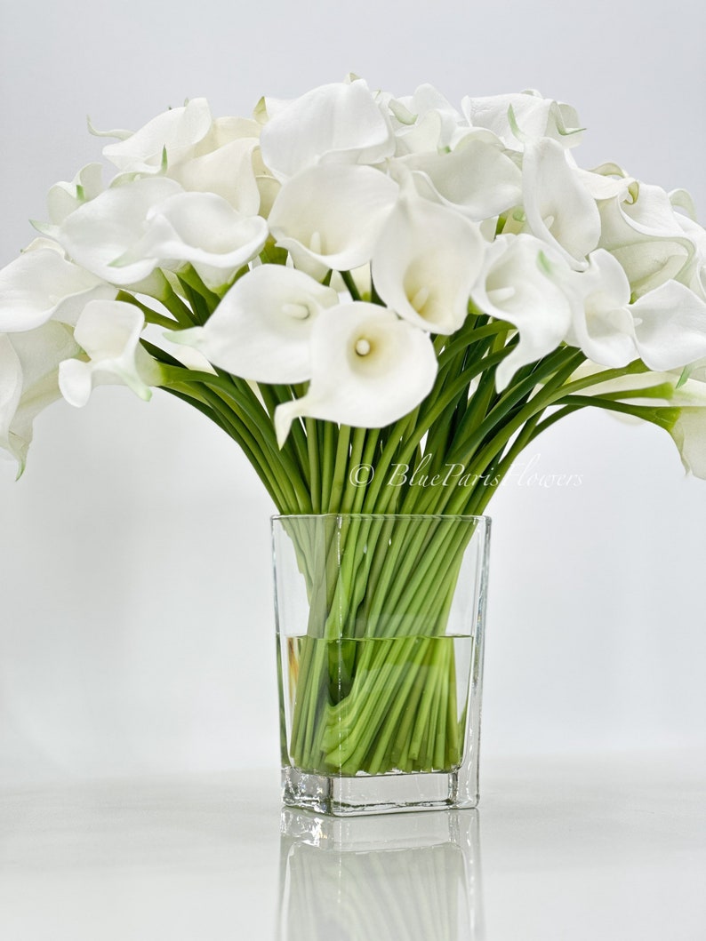 May include: A bouquet of white calla lilies arranged in a clear glass vase. The lilies are in full bloom and have long green stems.