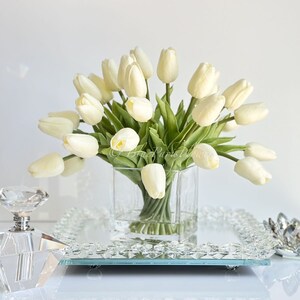 May include: A bouquet of white artificial tulips arranged in a clear glass vase. The vase is sitting on a mirrored tray with a crystal decorative piece.