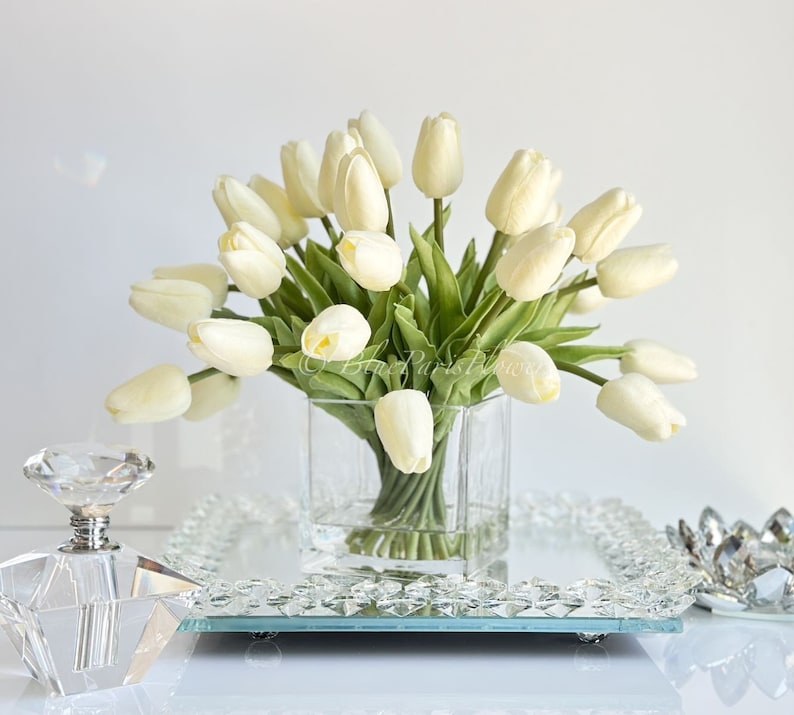 May include: A bouquet of white artificial tulips arranged in a clear glass vase. The vase is sitting on a mirrored tray with a crystal decorative piece.