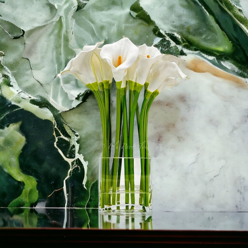 May include: A bouquet of white calla lilies arranged in a clear glass vase. The lilies are in a simple, modern style and are set against a green and white marble background.