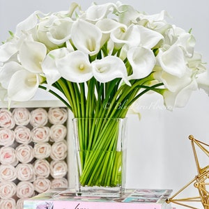 May include: A bouquet of white calla lilies in a clear glass vase, sitting on a stack of books with pink covers. The books have titles that include "love Manuela" and "Confetti Cakes".