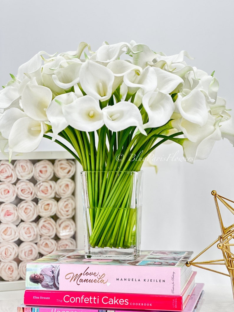 May include: A bouquet of white calla lilies in a clear glass vase, sitting on a stack of books with pink covers. The books have titles that include "love Manuela" and "Confetti Cakes".