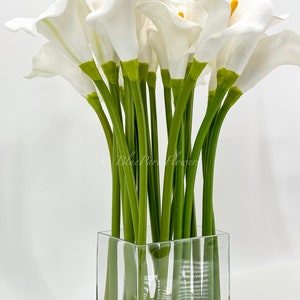 May include: A bouquet of white calla lilies arranged in a clear glass cube vase. The stems are green and the flowers are arranged in a symmetrical pattern.
